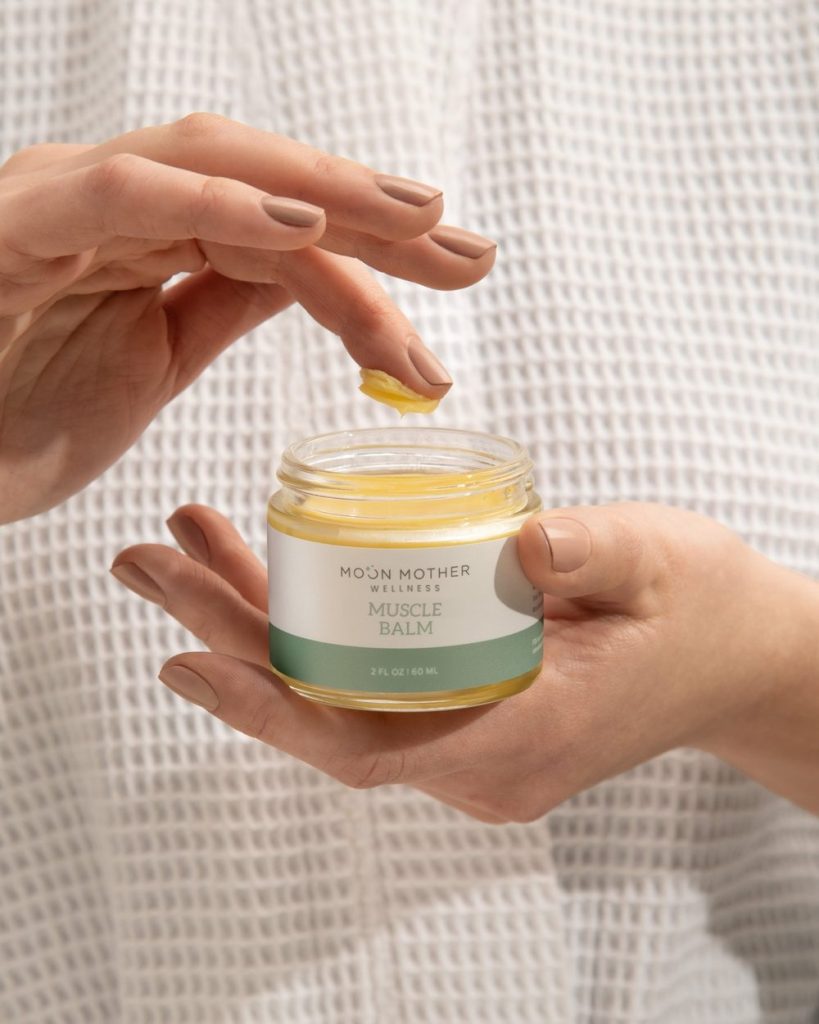 Hands scooping Muscle Balm from an open 2 fl oz jar